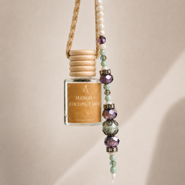 A small glass diffuser bottle labeled 'MANGO + COCONUT MILK' hangs from a woven cord with a beaded tassel. The bottle has a light wood cap and a warm tan label, while the hanging beads include white, mint green, and purple faceted stones with silver-toned accents, set against a soft beige background.