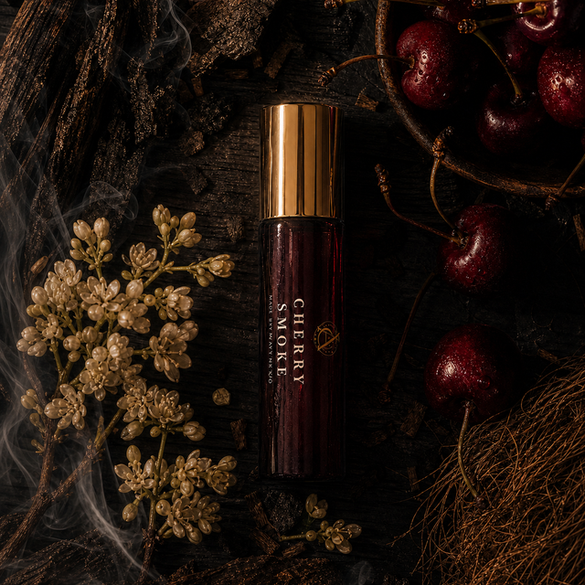 A dark amber glass bottle labeled 'CHERRY SMOKE' with a metallic gold cap, styled on a dark rustic surface with dried flowers, smoke, and fresh red cherries.