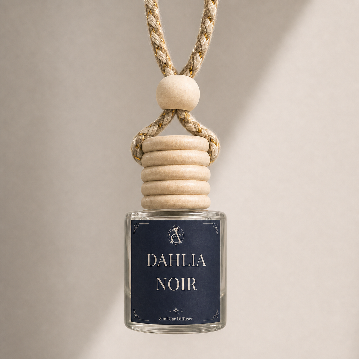 A glass car diffuser bottle with a navy blue label reading 'DAHLIA NOIR' and a light wood cap, suspended from a braided beige rope with a wooden bead, against a warm beige background.